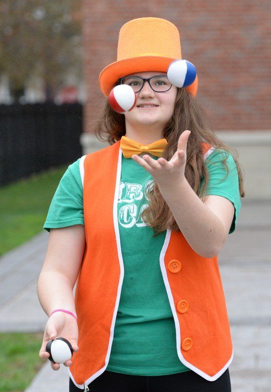 A performer with glasses in a green t-shirt, with an orange vest, hat, and bowtie, is juggling three small white balls